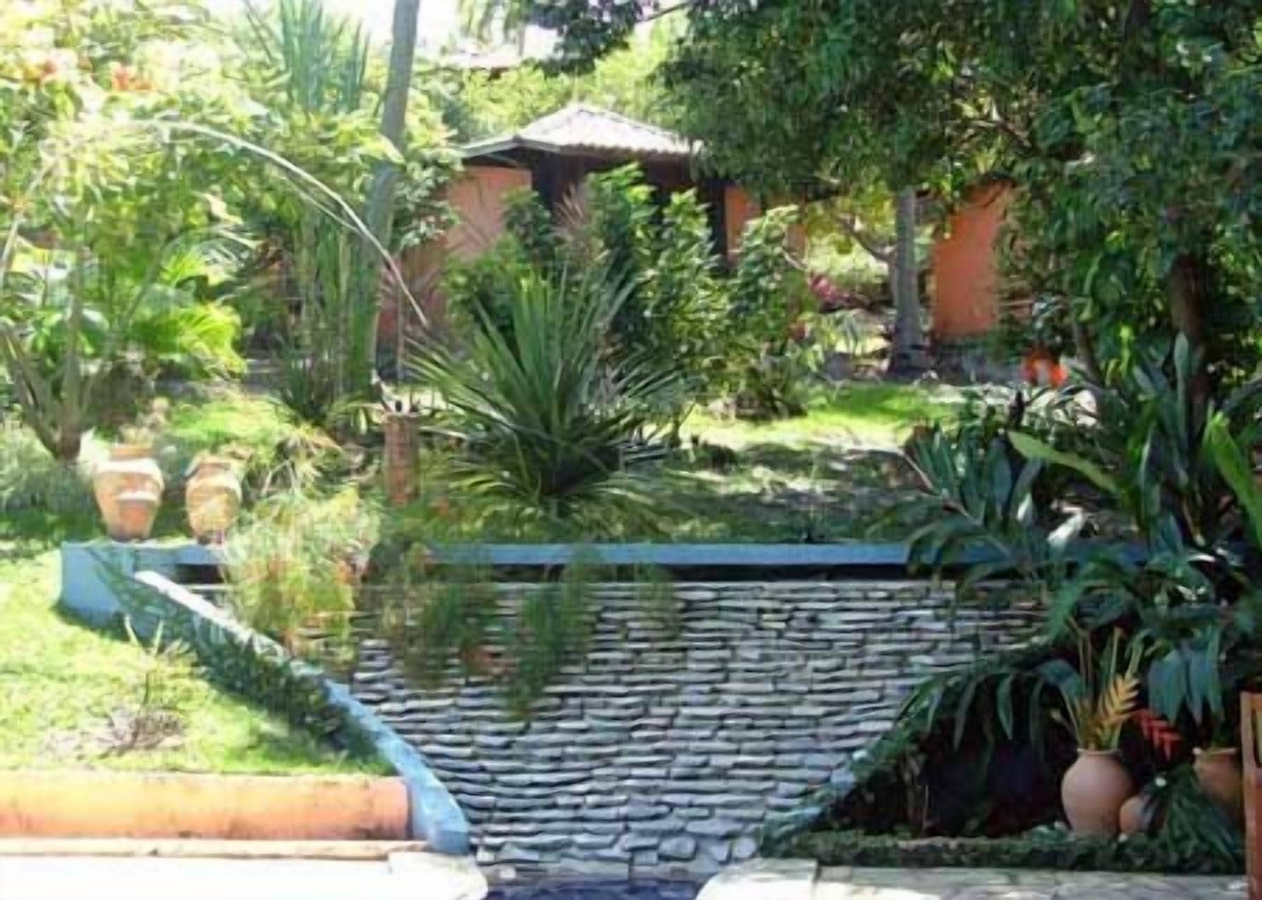 Un jardín con muro de piedra, diversas plantas y una pequeña fuente de agua.