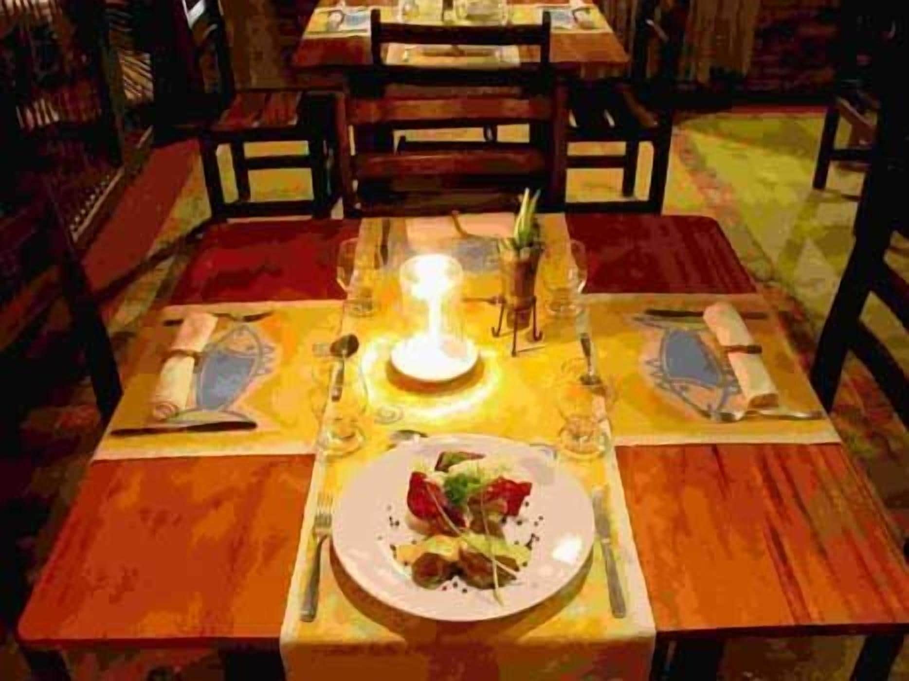 Mesa de comedor lista para dos personas, con una vela, platos, cubiertos y un vaso de agua.