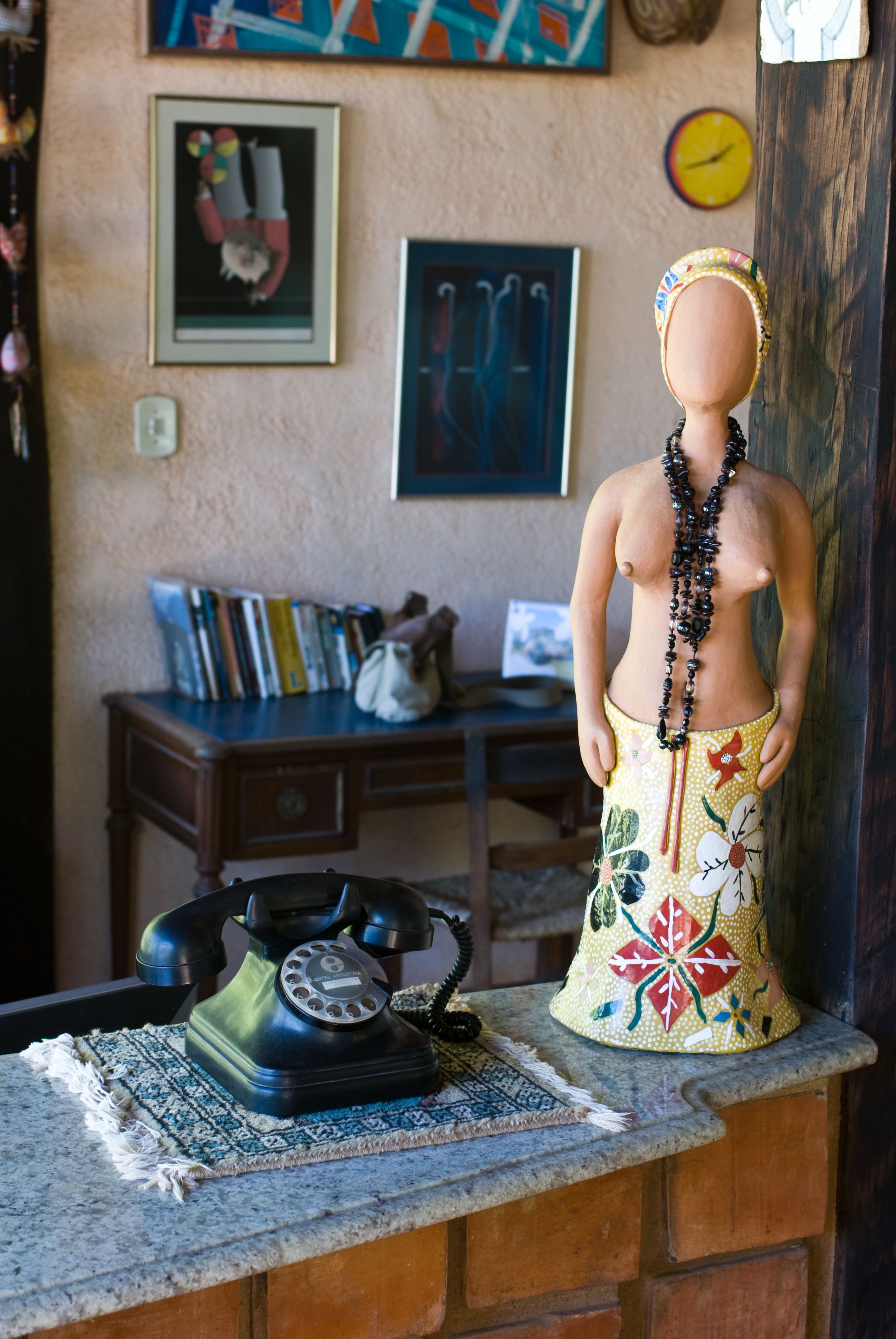 A mannequin with a floral skirt and a beaded necklace, a rotary phone, and a bookshelf with books and a framed picture in the background.