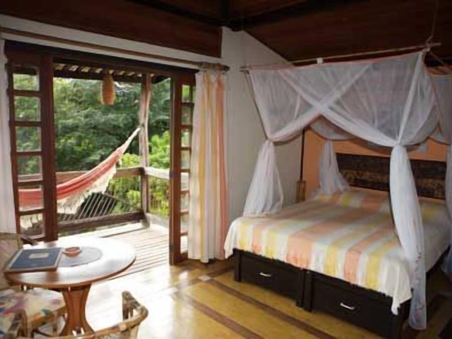 A bedroom with a canopy bed, a small table with a chair, and a view of greenery through a glass door.