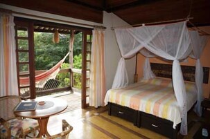 A bedroom with a canopy bed, a small table with a chair, and a view of greenery through a glass door.