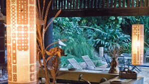 Una zona de bar con salpicadero de azulejos, un lavamanos y un jarrón con plantas secas. Hay sillas de mimbre y techo de madera.