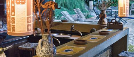 Un coin bar avec un mur recouvert de carreaux, un évier et un vase contenant des plantes séchées. On y trouve des chaises en osier et un plafond en bois.