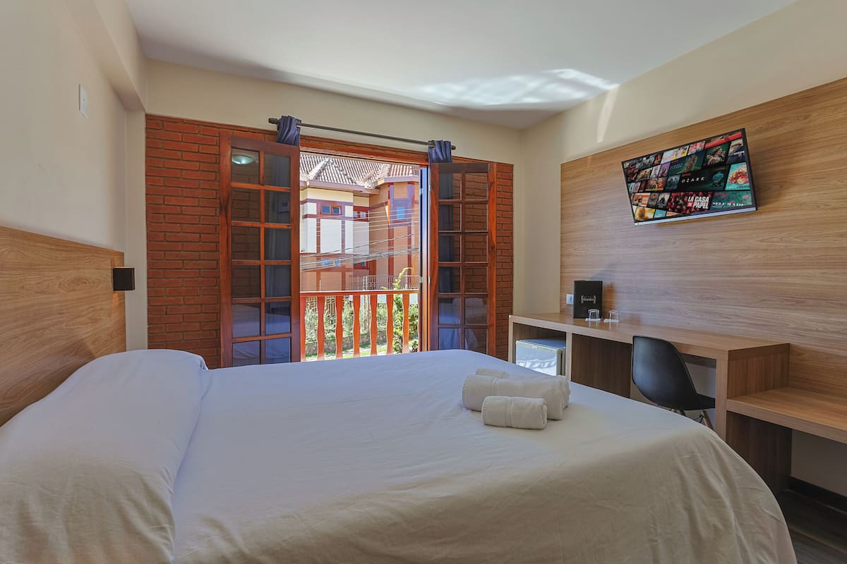 A hotel room with a large bed, a desk, a TV mounted on the wall, and a balcony with a view of the outside.