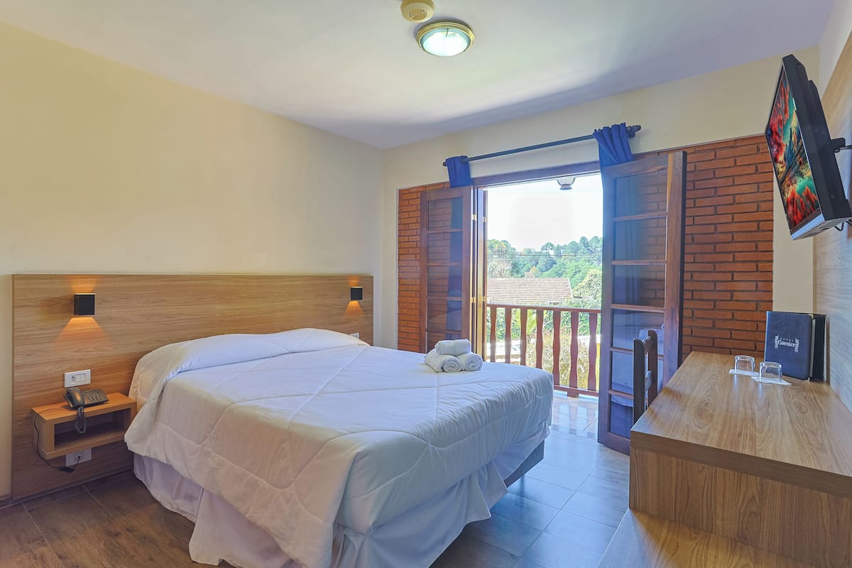 A hotel room with a large bed, a wooden nightstand with a phone, a desk with a TV, and a balcony with a view of greenery.