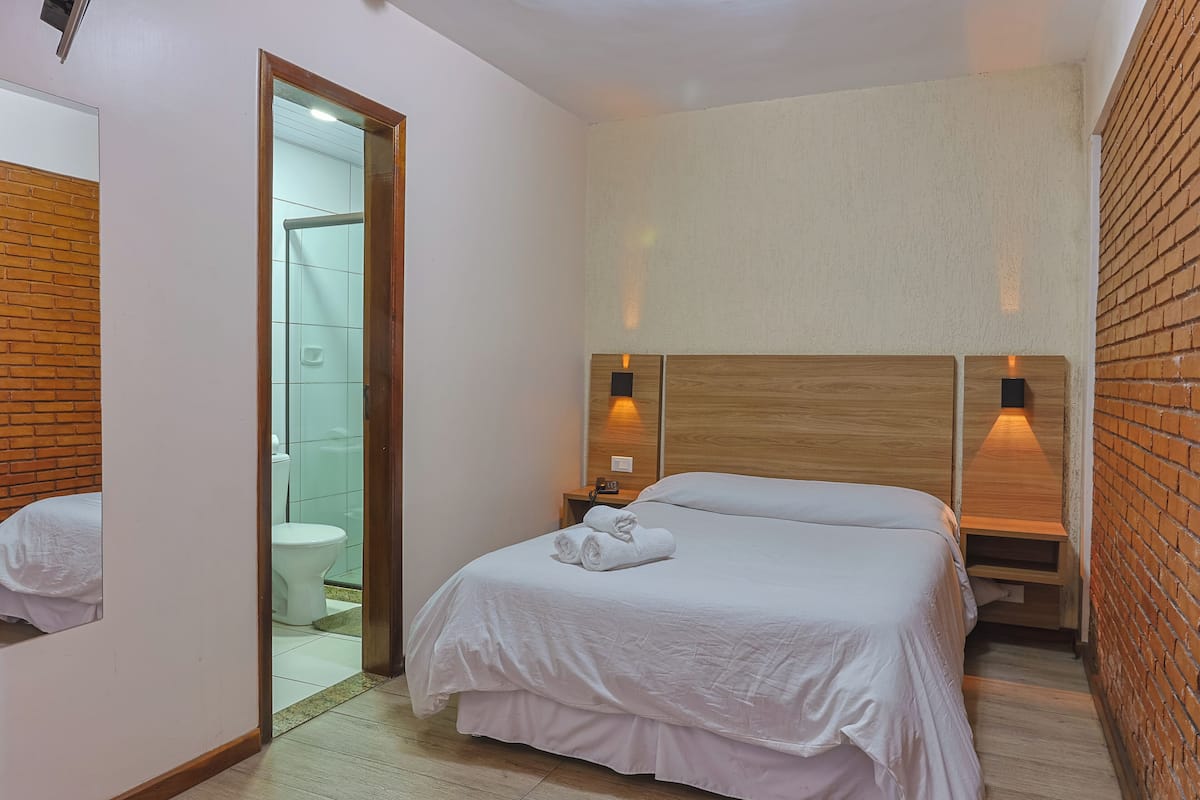 A hotel room with a wooden headboard, a bed with white linens, a nightstand with a lamp, and a view of the bathroom through an open door.