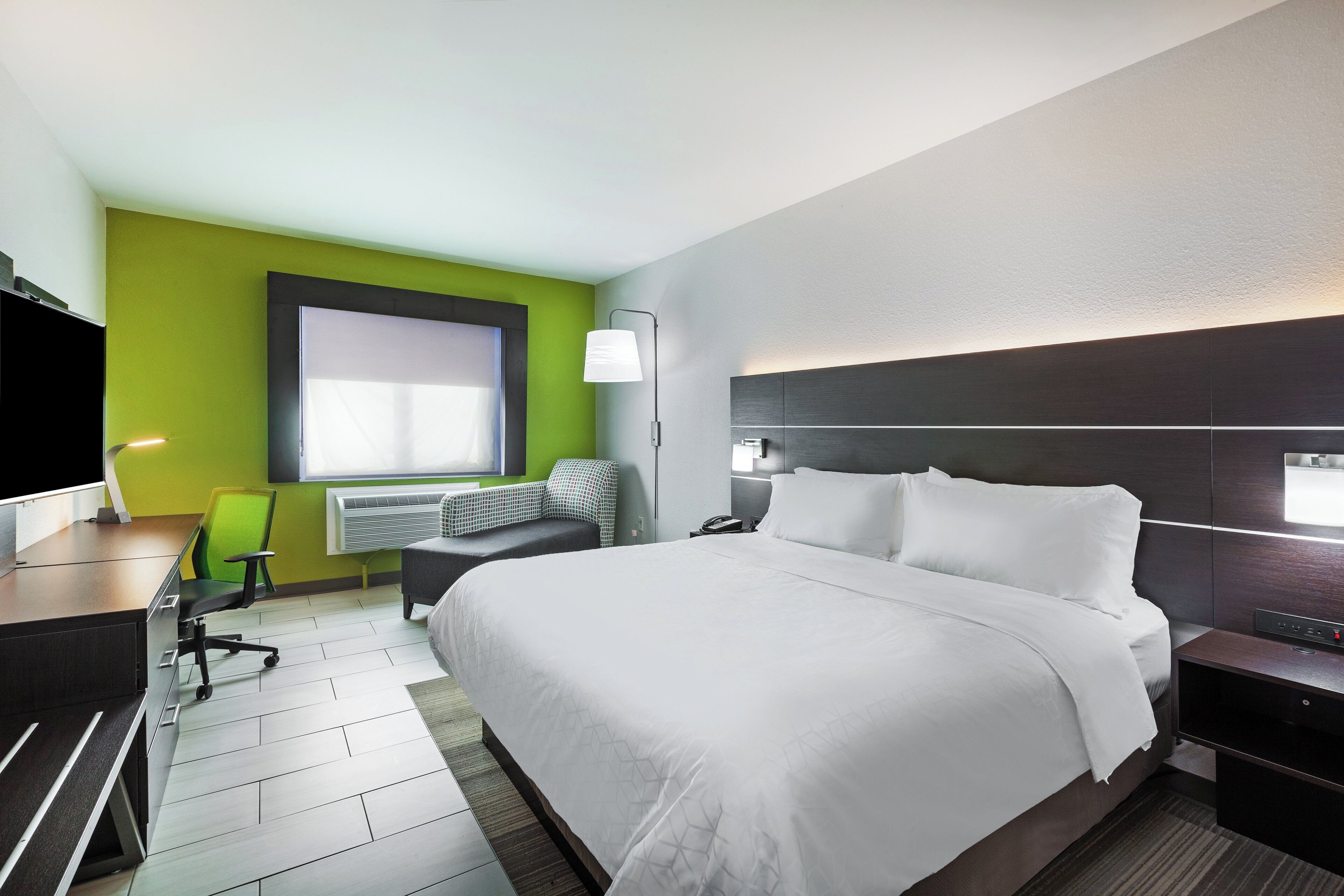 A hotel room with a large bed, a desk with a chair, a standing lamp, and a window with blinds.
