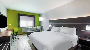 A hotel room with a large bed, a desk with a chair, a standing lamp, and a window with blinds.