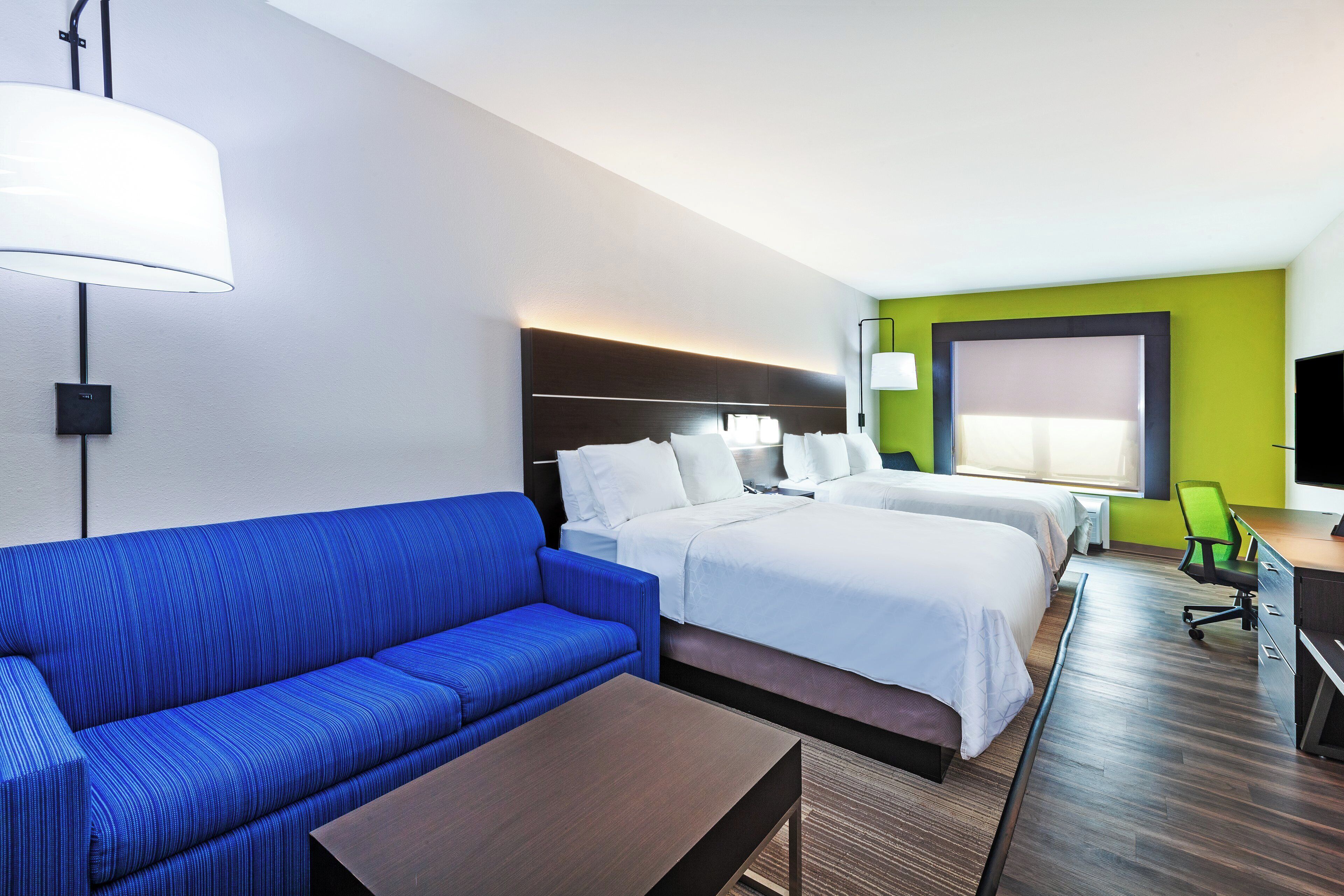 Habitación de hotel con sofá azul, dos camas, un escritorio y un ventanal grande.