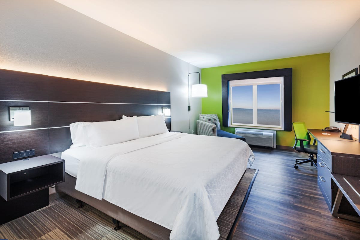 A modern hotel room with a large bed, bedside tables, a desk with a chair, a window with a view, and a flat-screen TV.