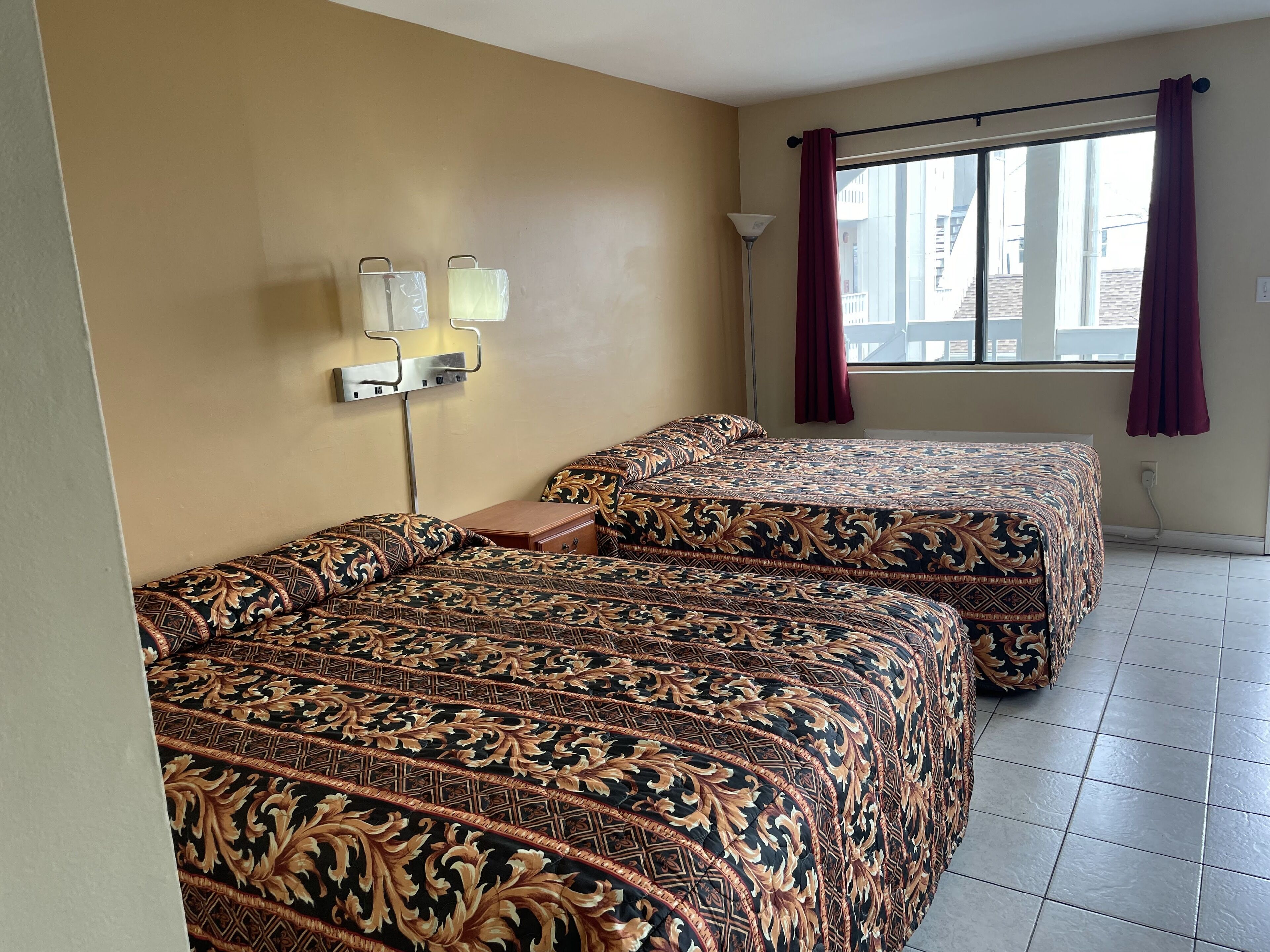 A hotel room with two beds, a nightstand, a lamp, and a window with curtains.