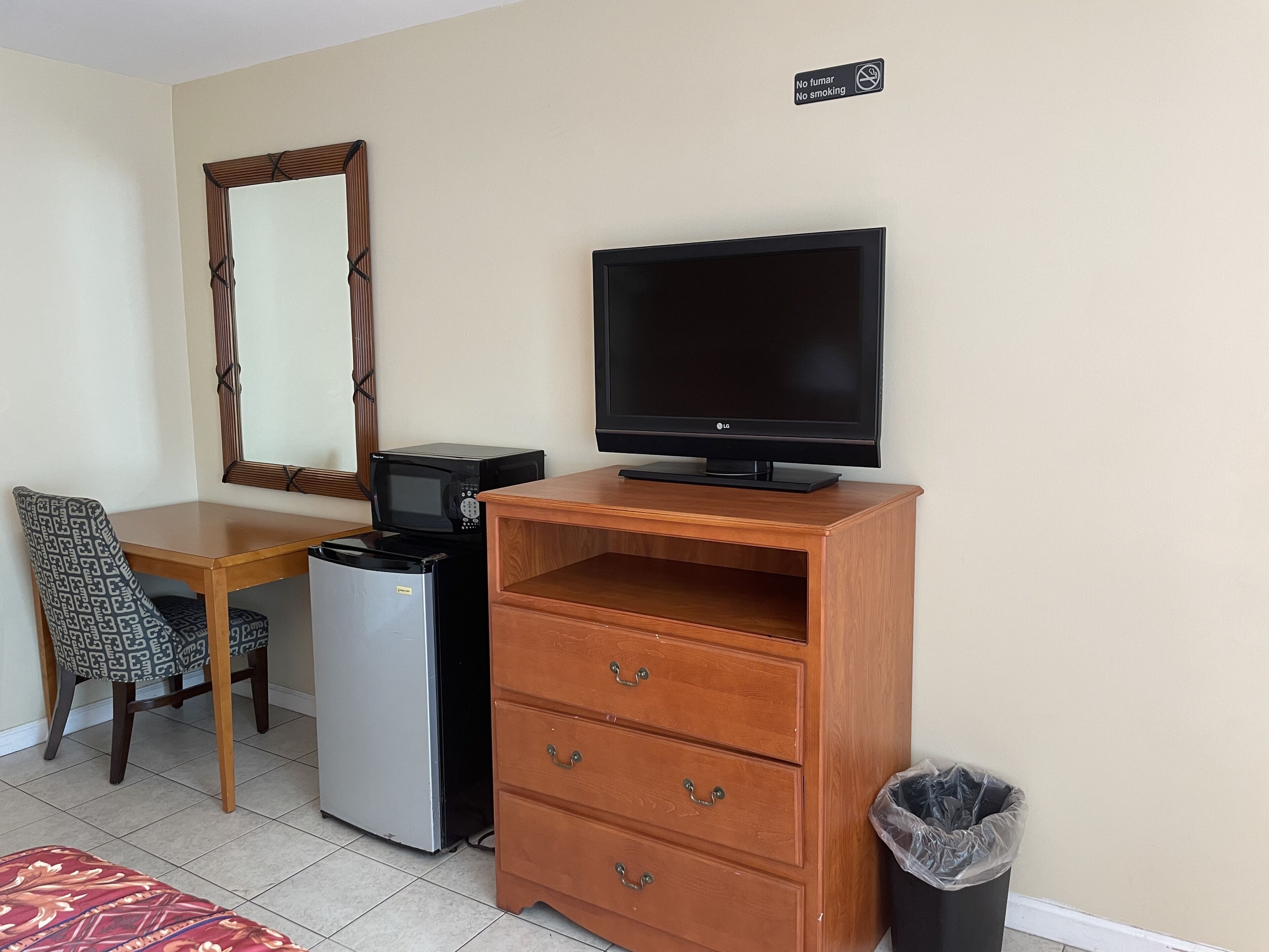 A small room with a wooden desk, a microwave, a refrigerator, a television on a wooden stand, a chair, a mirror, and a trash bin.