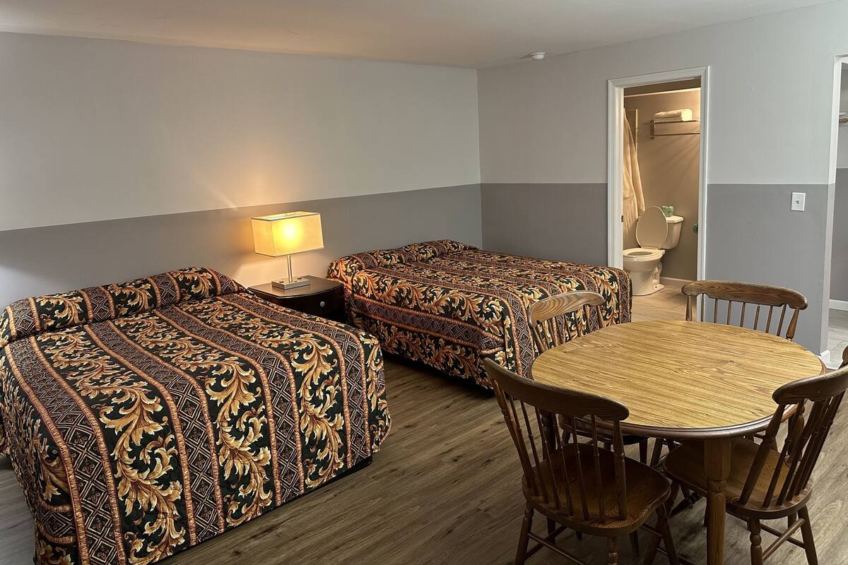 A room with two beds, a table, and chairs. There is a lamp on a nightstand.