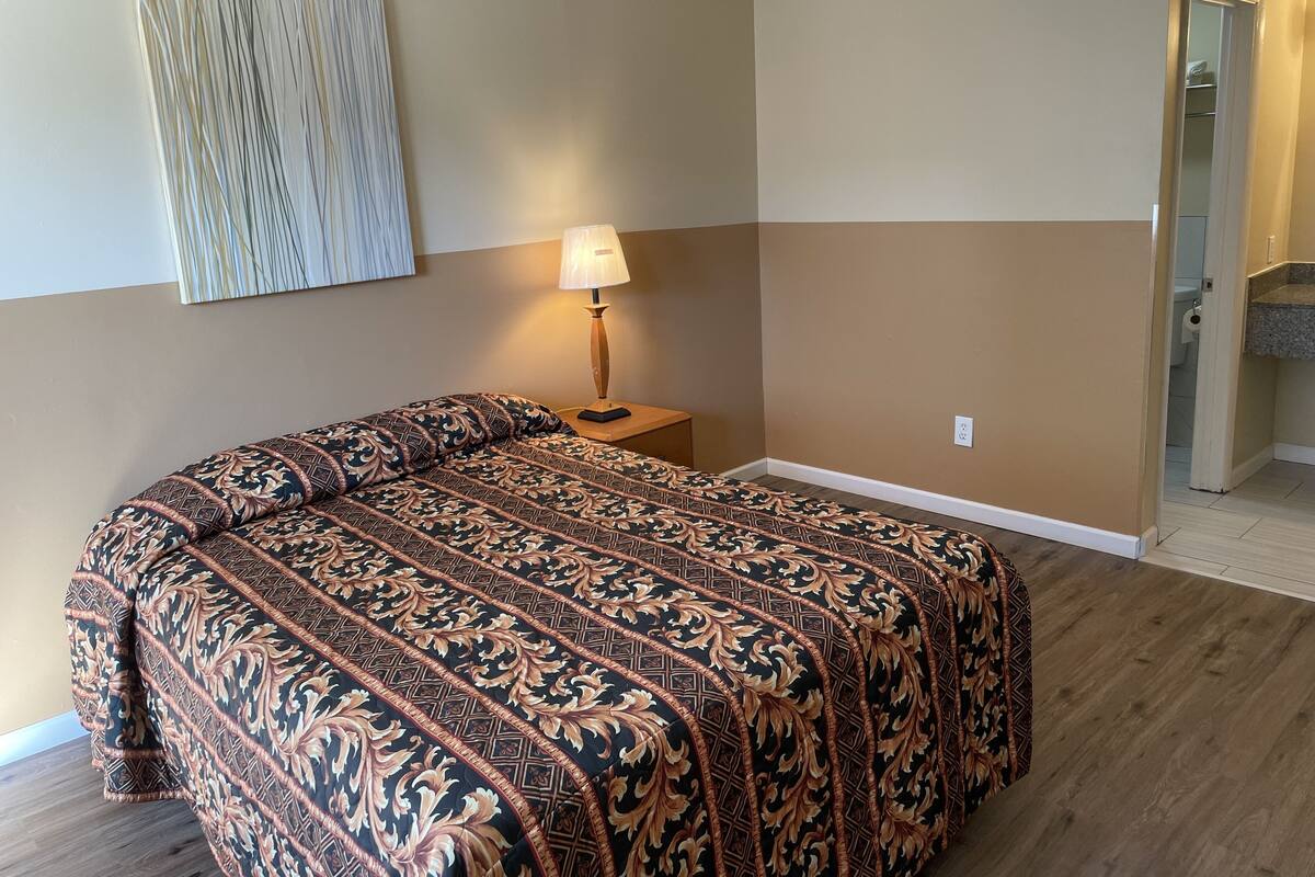 A hotel room with a large bed, a nightstand with a lamp, a wall-mounted artwork, and a door leading to a bathroom.