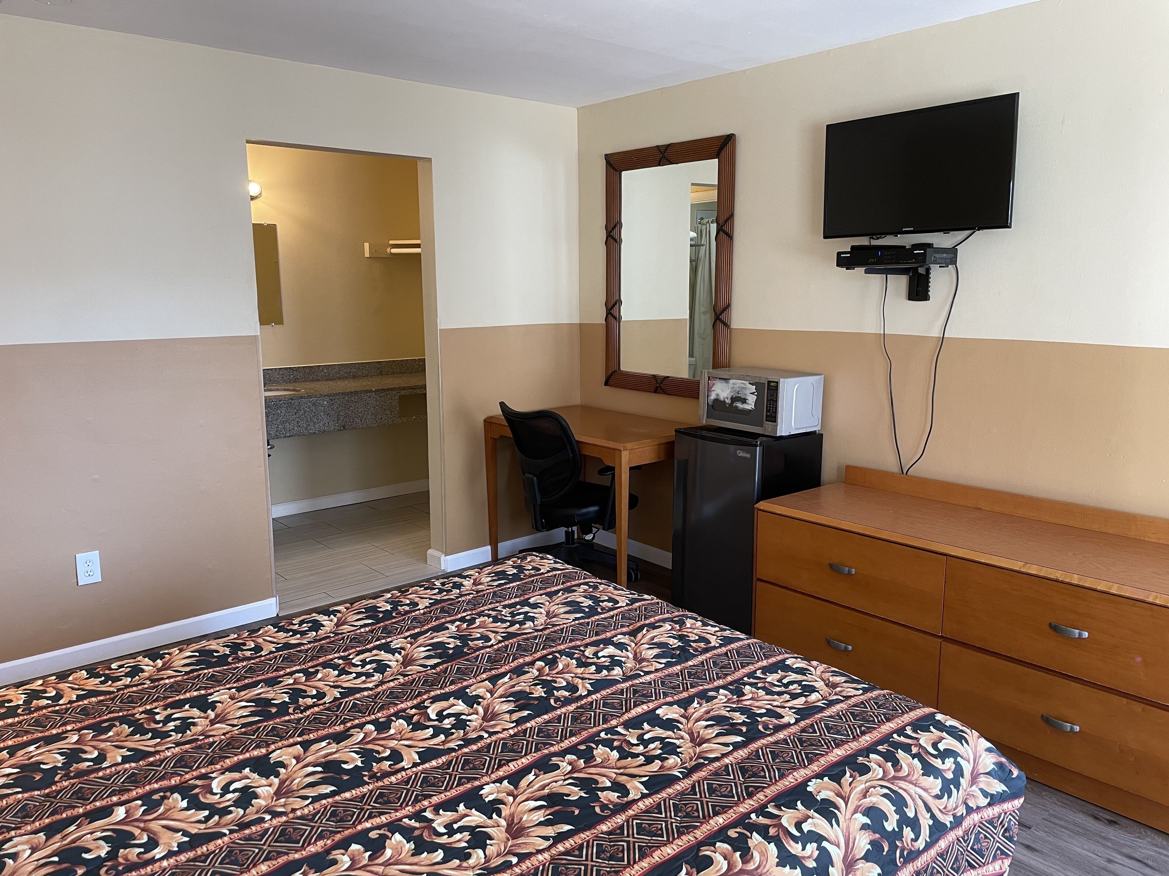 A hotel room with a bed, a desk, a refrigerator, a dresser, a television, and a mirror.