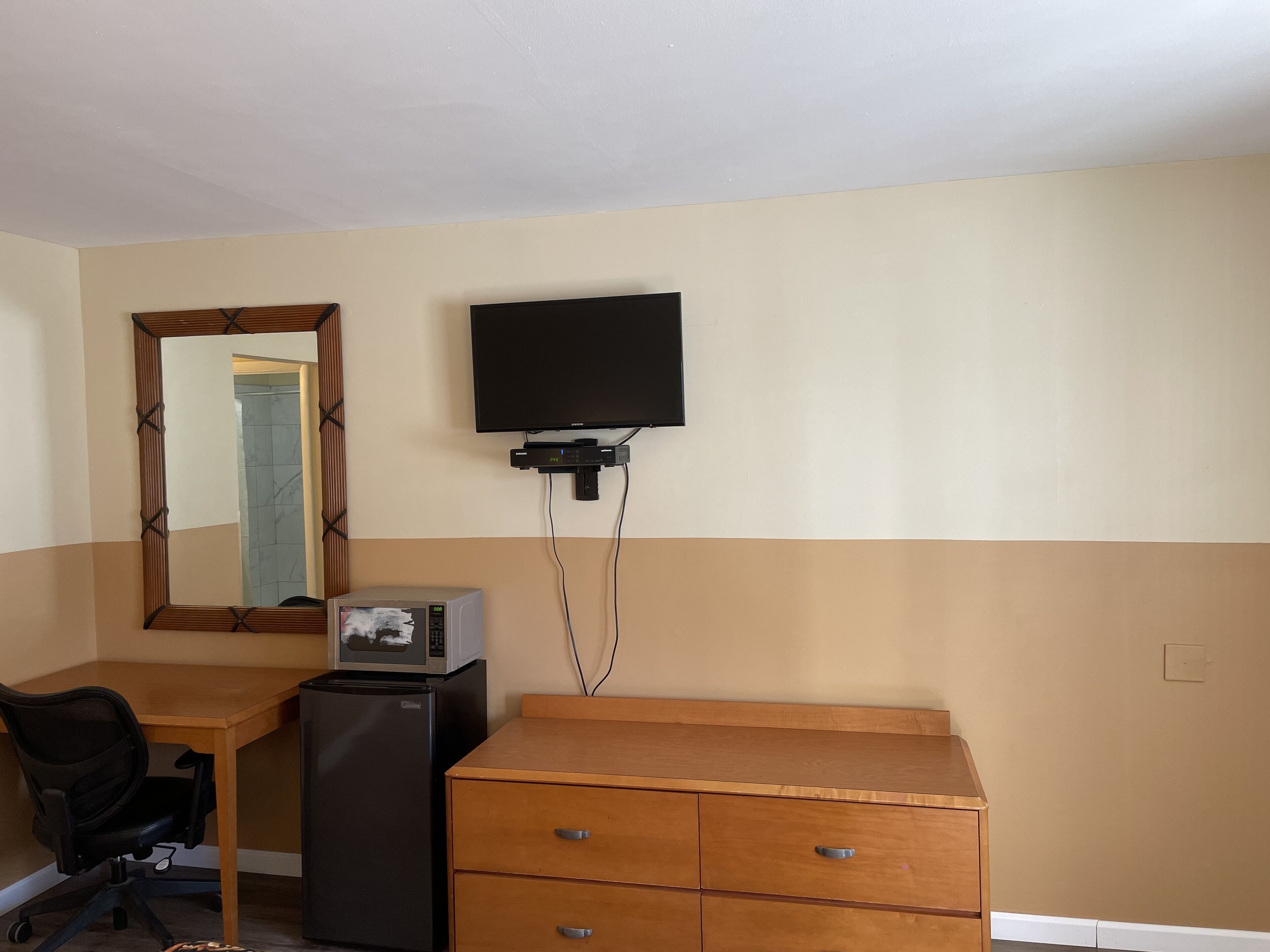 A hotel room with a wooden desk, a black office chair, a refrigerator, a wooden dresser with drawers, a flat-screen TV mounted on the wall, and a mirror with a wooden frame.