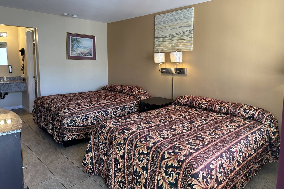 A hotel room with two beds, a small desk, a mirror, a painting, and a wall-mounted lamp.