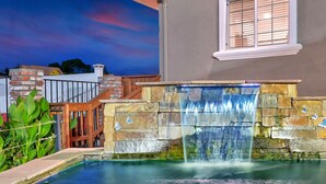 A stone waterfall feature in a backyard with a pool, surrounded by a wooden deck and a brick wall. - Traveler's Inn (Williams)