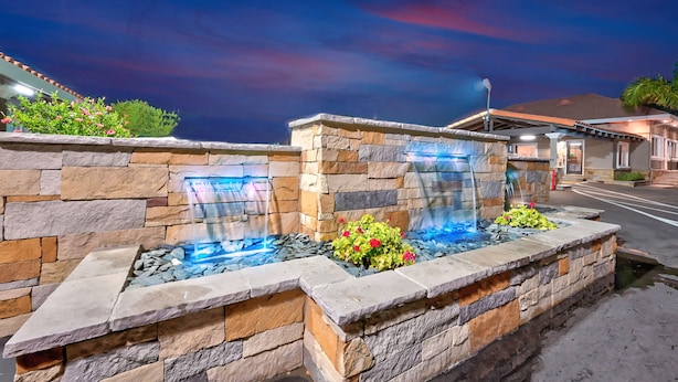 A stone water feature with a waterfall, surrounded by a stone wall and plants, set against a twilight sky. - Traveler's Inn (Williams)