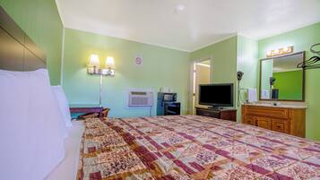 A hotel room with a large bed, a television, a vanity with a mirror, and a wall-mounted air conditioner.