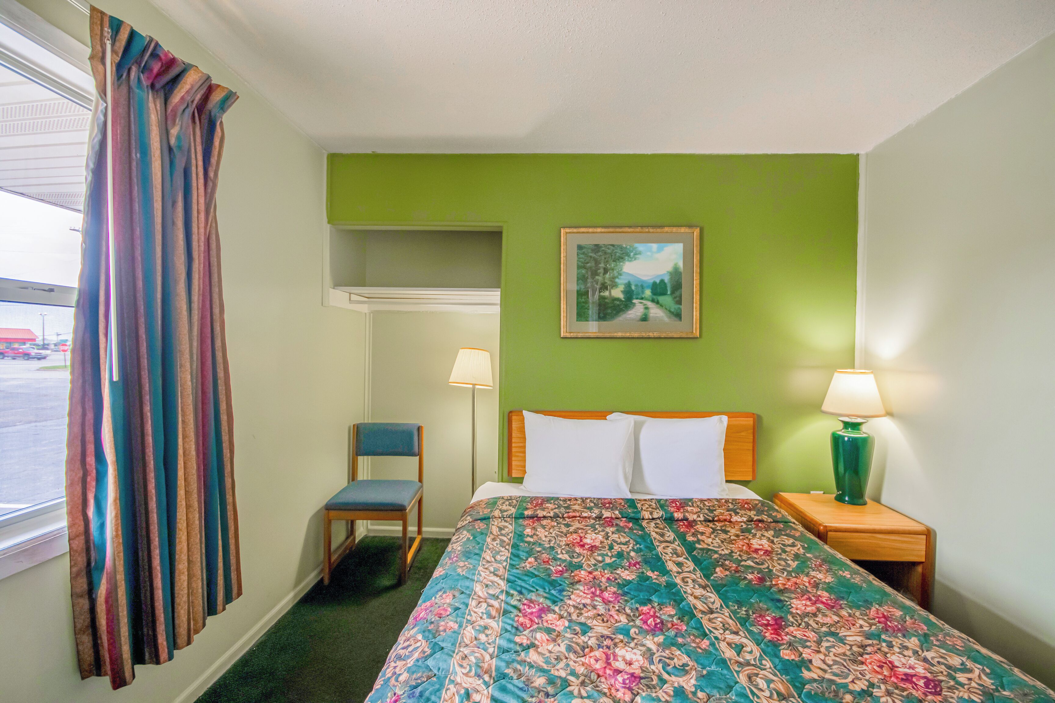 Habitación de hotel con cama, una silla, mesita de noche con lámpara, un cuadro en la pared y ventana con cortinas.
