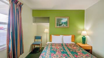 Habitación de hotel con cama, una silla, mesita de noche con lámpara, un cuadro en la pared y ventana con cortinas.
