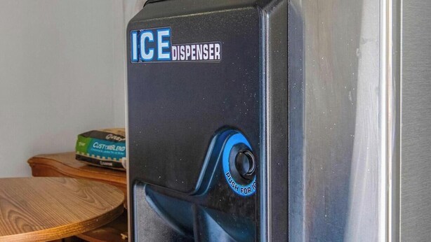 A stainless steel ice dispenser with a blue button labeled 'PUSH FOR ICE' in a kitchen setting.