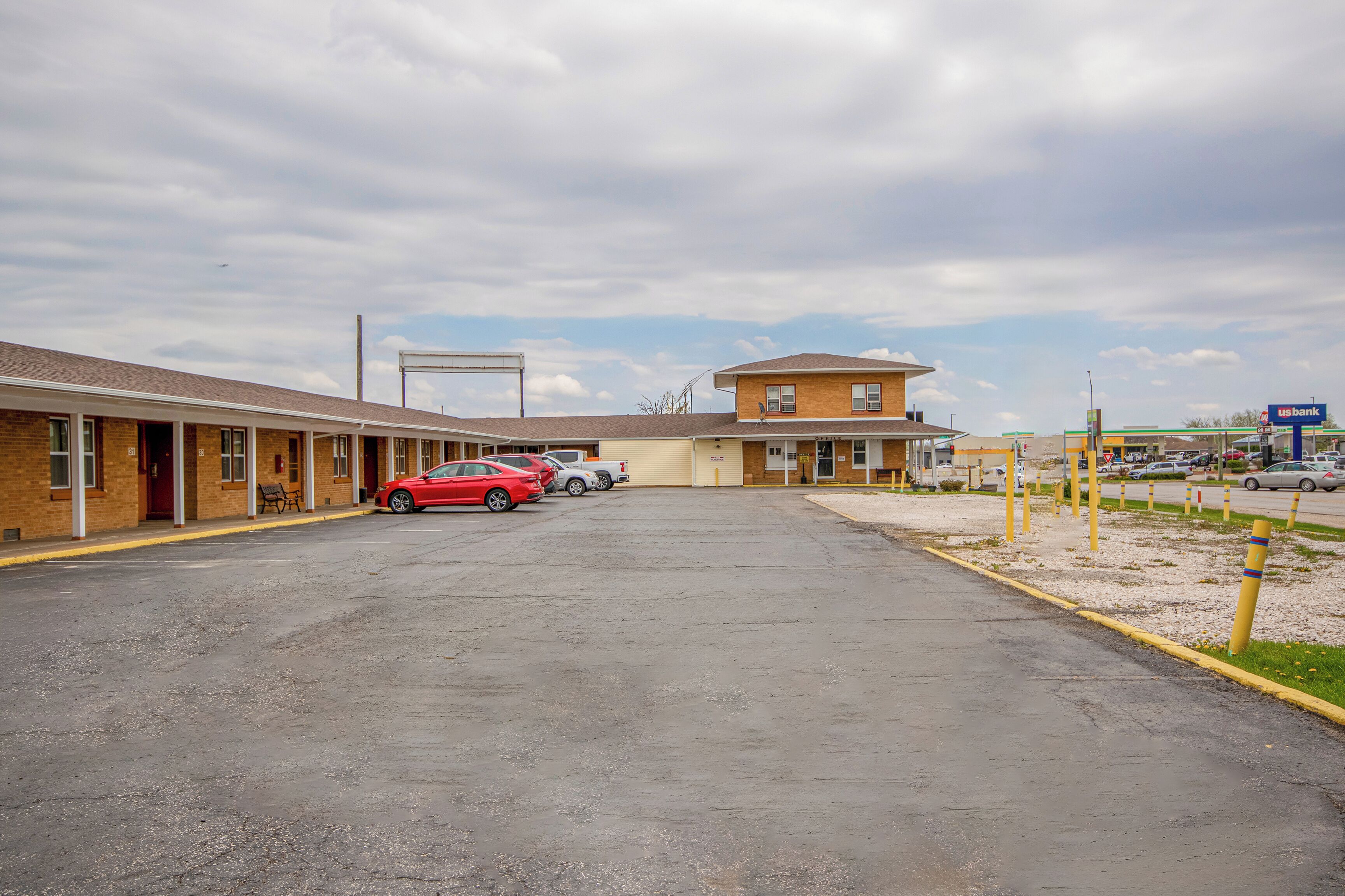 Un motel con estacionamiento, un coche rojo y un letrero de banco al fondo.