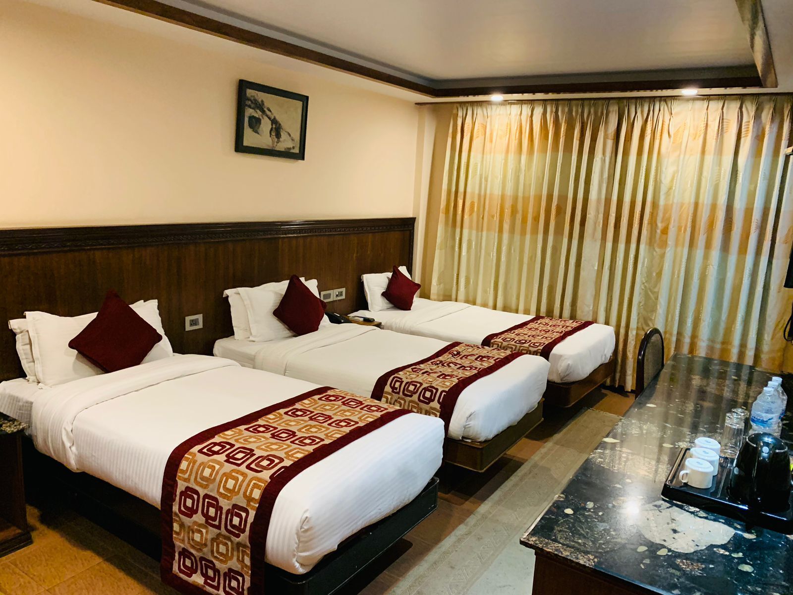 A hotel room with three beds, each with white bedding and patterned throw blankets. There is a large window with curtains, a bedside table with a tray of coffee cups and a water bottle, and a framed picture on the wall.