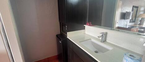 A bathroom with a white sink, dark cabinets, a black microwave, and a mirror above the sink.