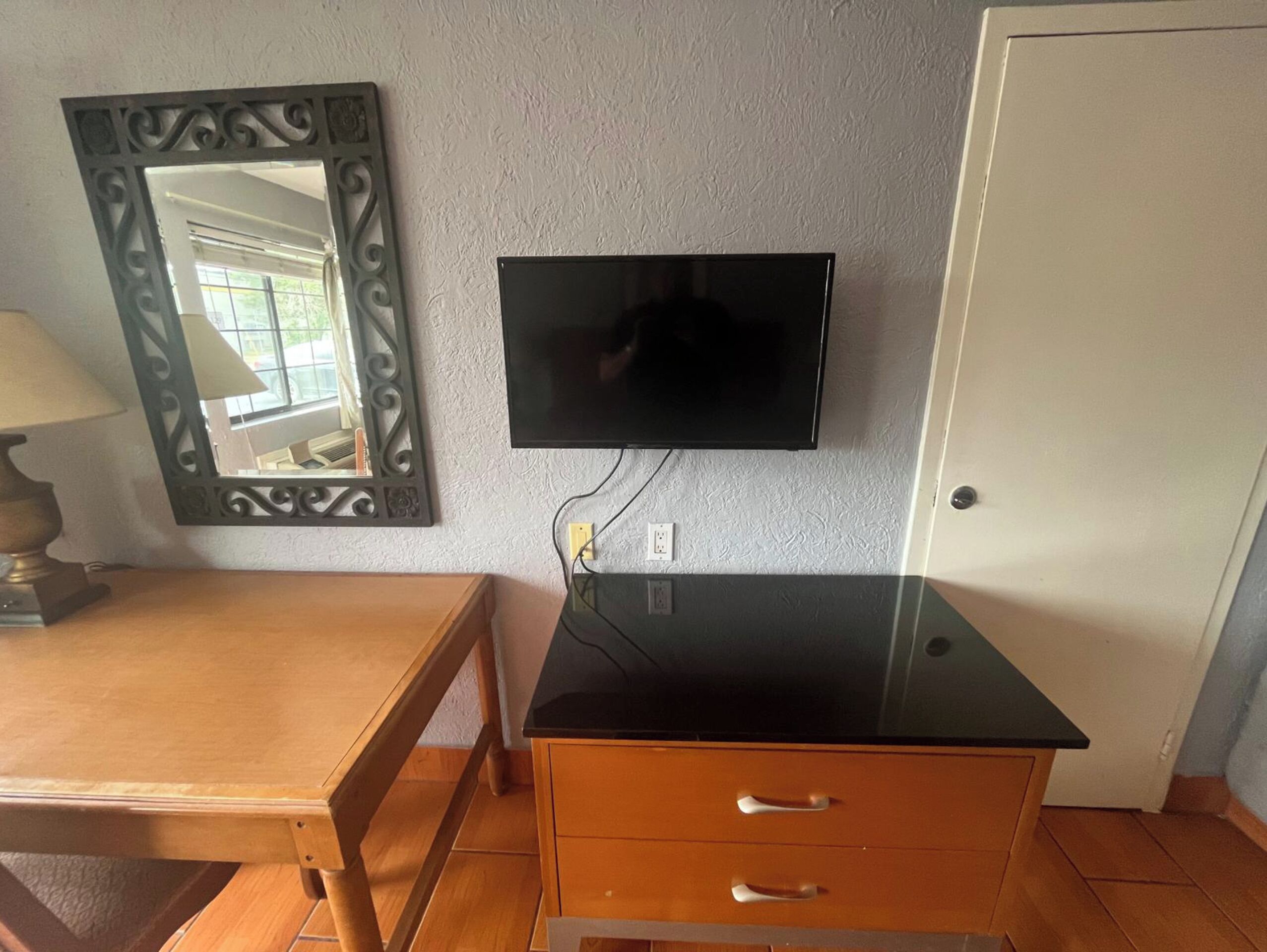 Une pièce avec un bureau en bois, un téléviseur noir fixé au mur, un miroir décoratif et une porte.