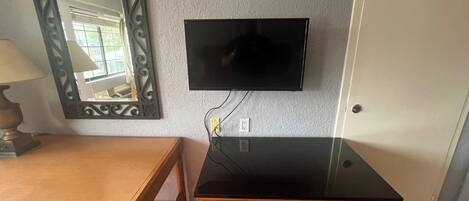 Une pièce avec un bureau en bois, un téléviseur noir fixé au mur, un miroir décoratif et une porte.
