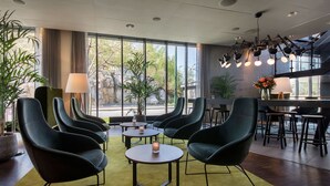 A modern lounge area with round tables and dark chairs, large windows with a view of greenery, and a bar area with stools.