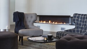 A modern living room with a fireplace, two armchairs, a coffee table with a newspaper and a candle, and a coat draped over one armchair.