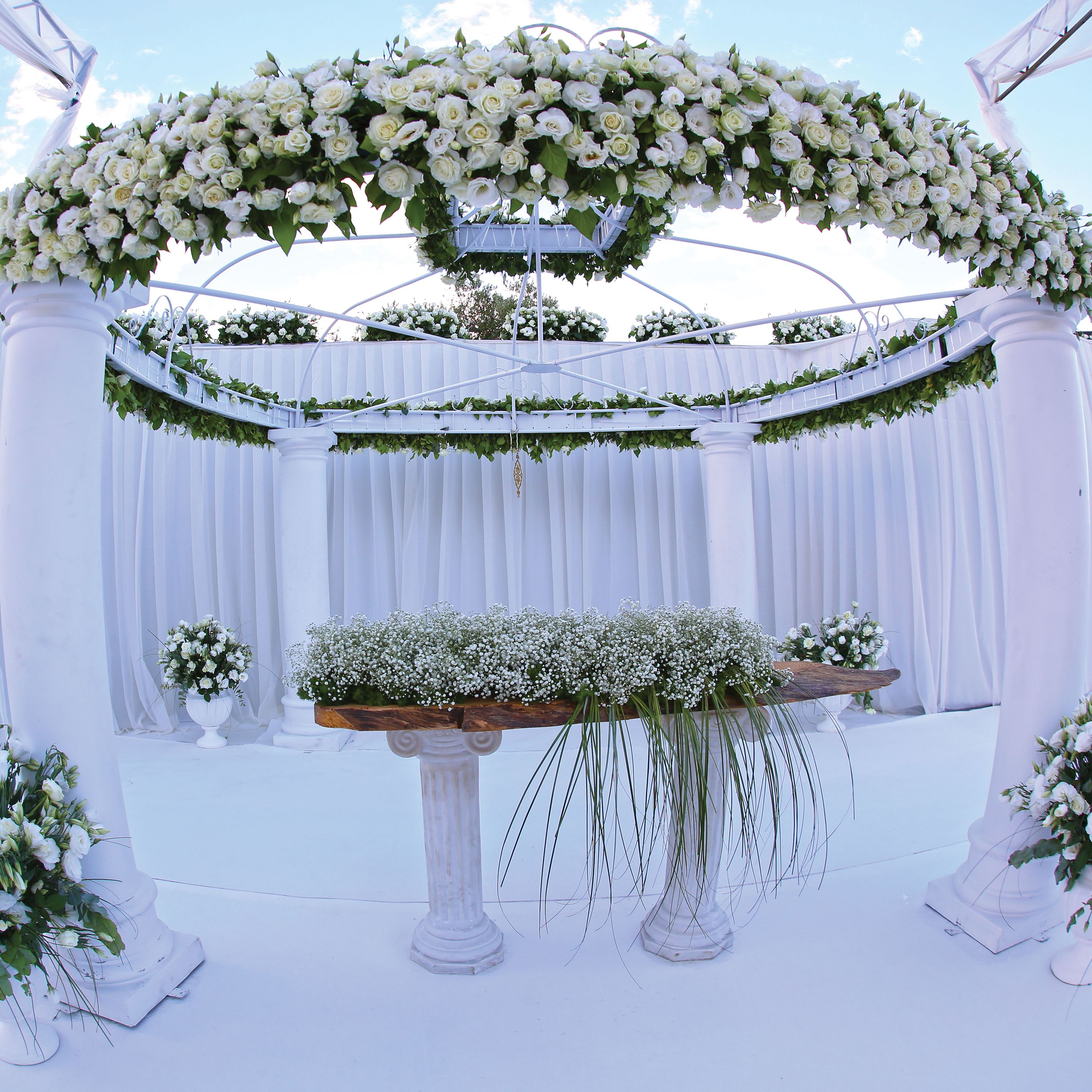 A white floral arch with a wooden platform at its center, surrounded by white columns and draped curtains.