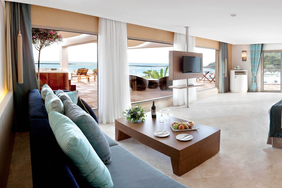 Ein modernes Hotelzimmer mit einem großen Bett, einer Couch, einem Couchtisch und einem Fernseher. Ein Balkon bietet Blick aufs Meer.