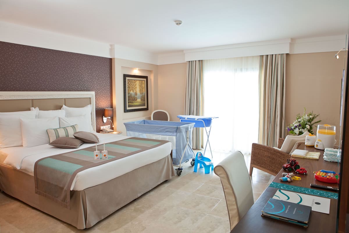 Ein Hotelzimmer mit einem großen Bett, einem Schreibtisch mit einem Laptop, einem Sessel, einem kleinen Tisch mit Spielzeug und einem Fenster mit Vorhängen.