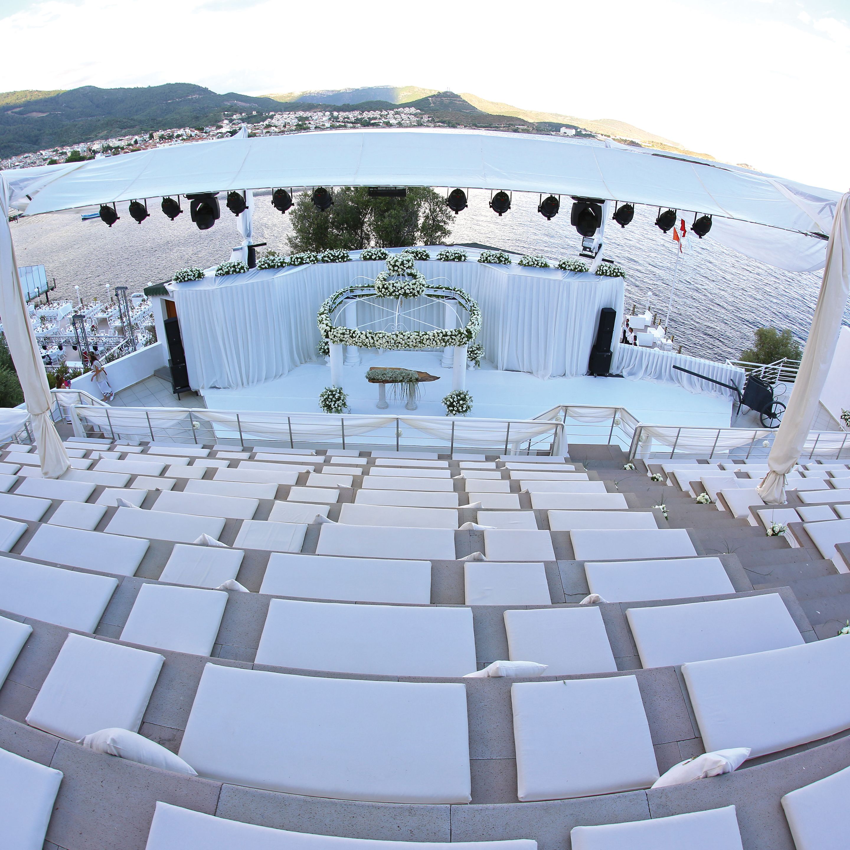 An outdoor amphitheater with tiered seating, a stage decorated with floral arrangements, and a backdrop of a coastal landscape.