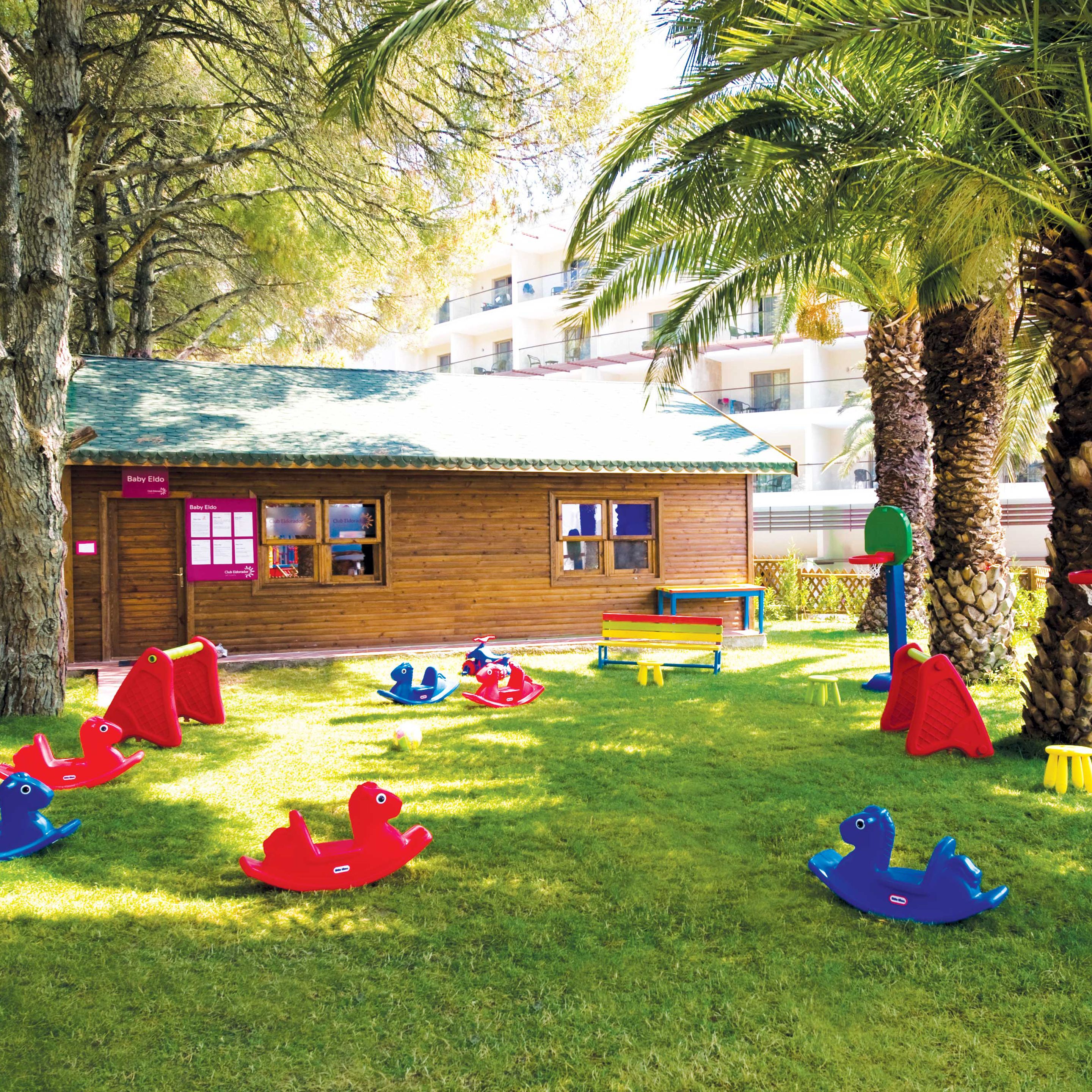A wooden building with a green roof, surrounded by palm trees and a grassy area with children's play equipment.