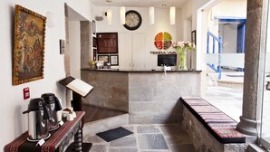 Una zona de recepción con piso de piedra, un reloj, un cuadro religioso enmarcado, una estación de café y vistas al exterior.