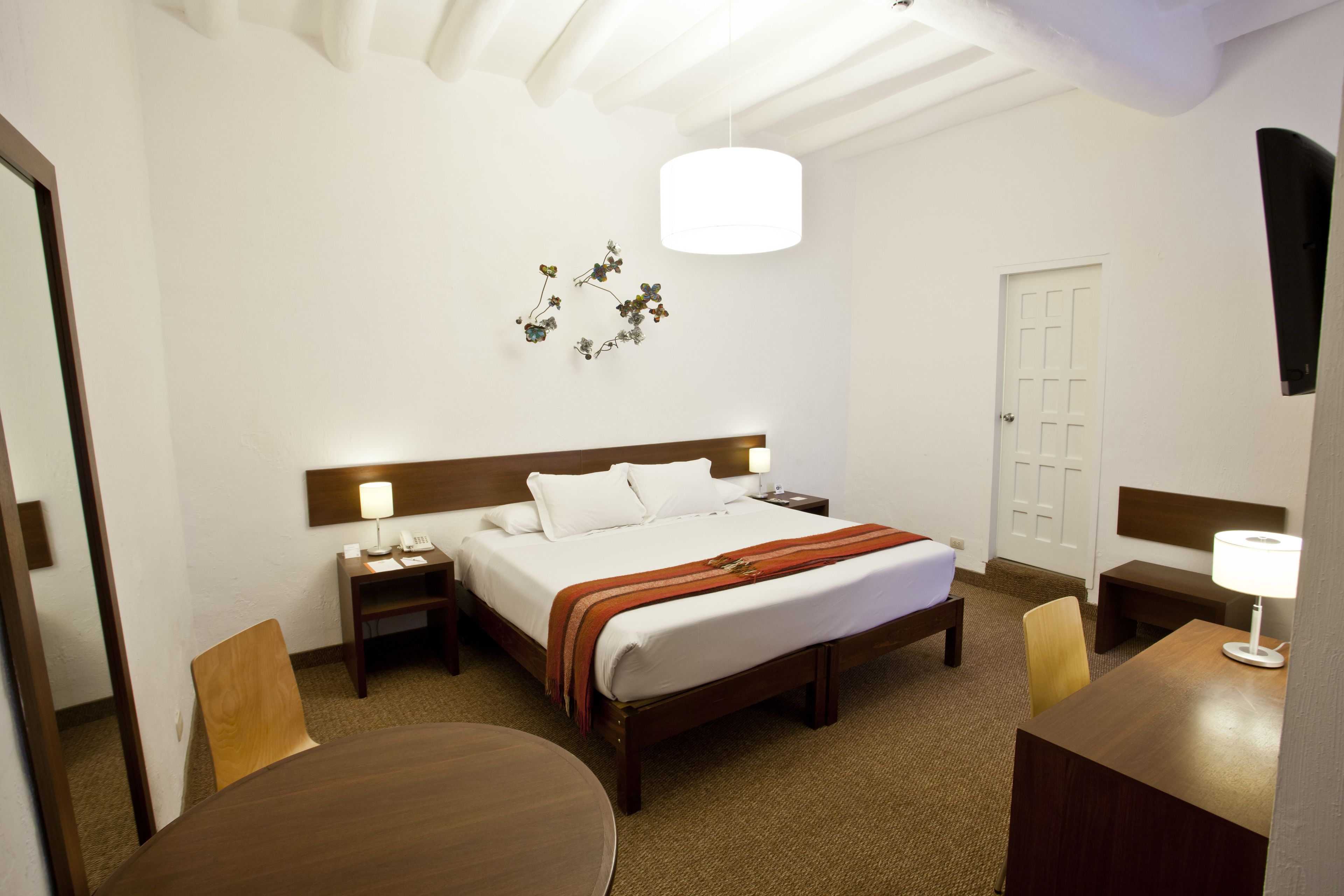 Habitación de hotel con cama, mesitas de noche, escritorio, sillas, una mesa redonda, lámpara colgante y una puerta.
