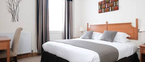 A hotel room with a large bed, a desk, a chair, a radiator, and a window with curtains.