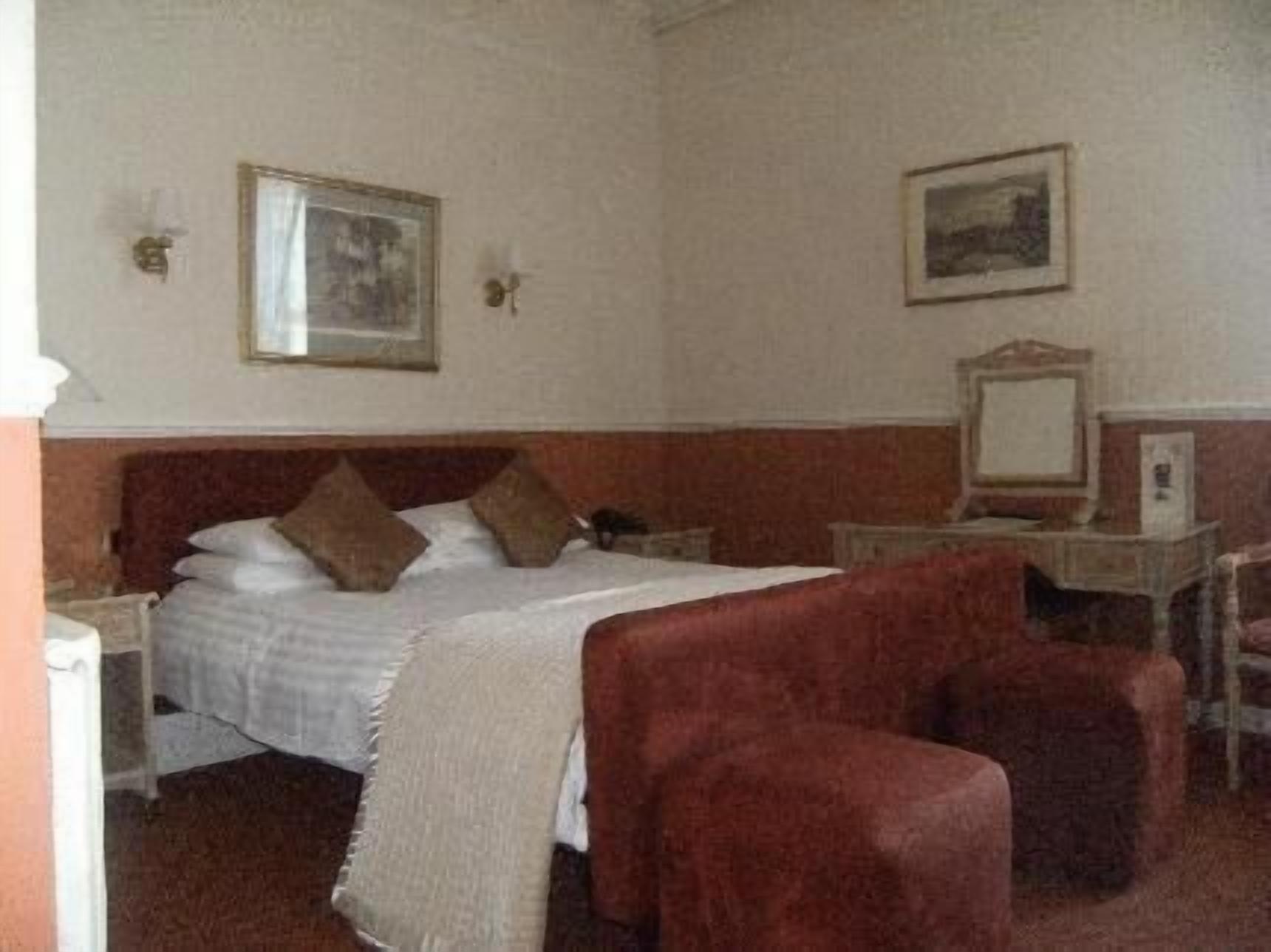 A hotel room with a bed, two chairs, a desk with a mirror, and two framed pictures on the wall.