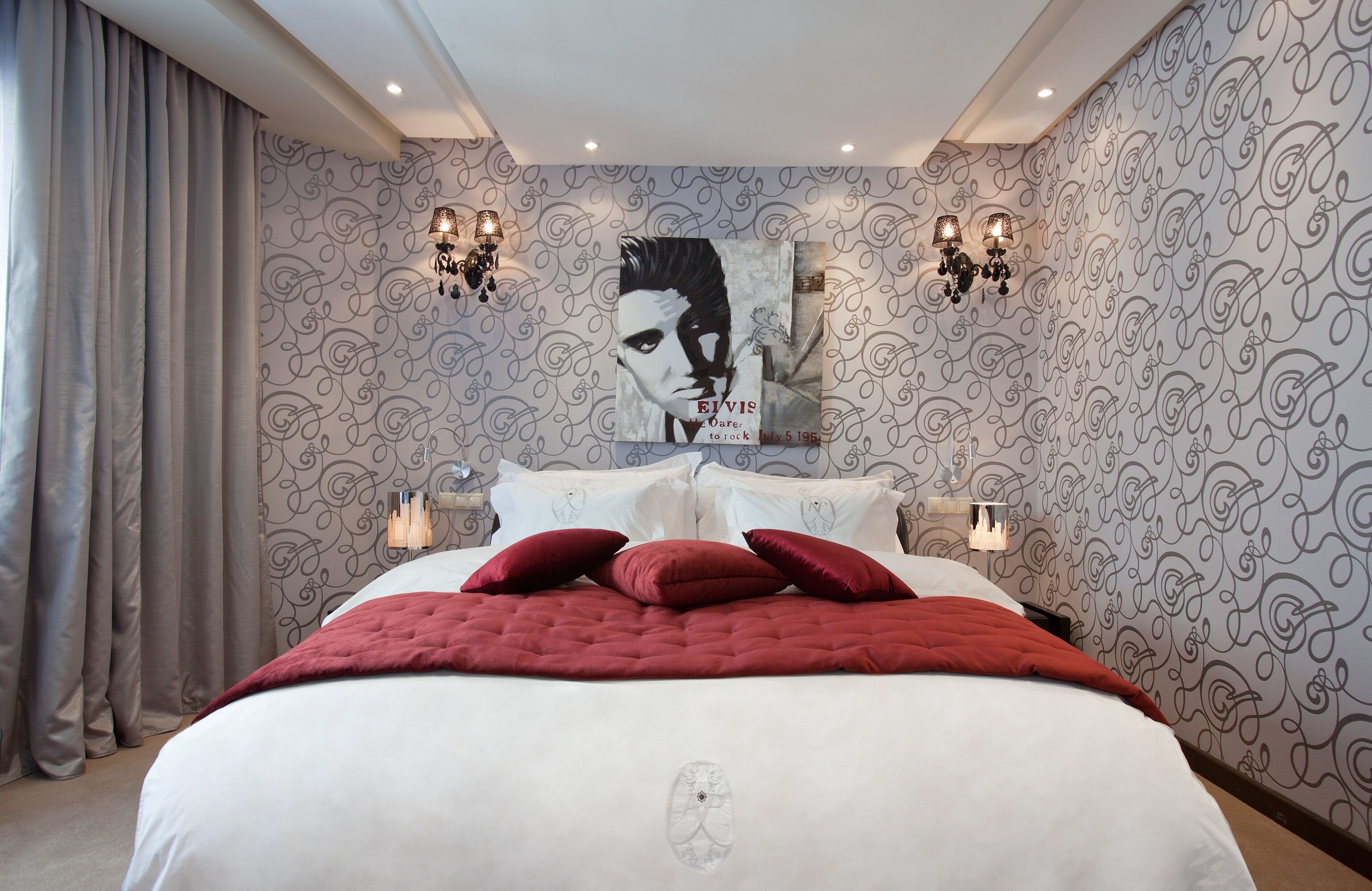 A bedroom with a large bed, patterned wallpaper, and wall sconces.