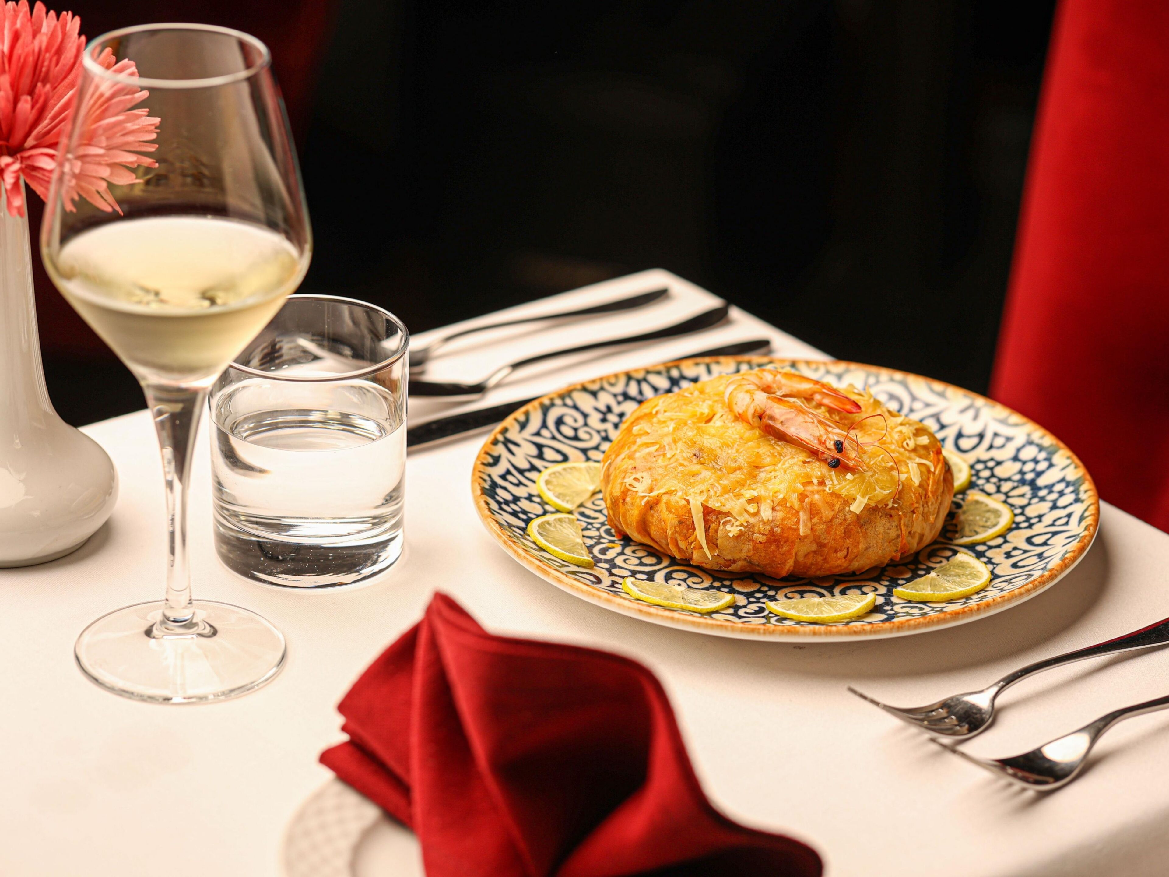 Une assiette avec une pâtisserie, un verre d’eau et un verre de vin blanc, posés sur une table recouverte d’une serviette rouge et agrémentée de couverts.