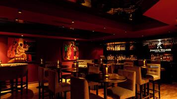 Una zona de bar con diversas opciones de asientos, incluyendo mesas redondas y taburetes de bar. Se observan varias botellas y vasos en la barra.