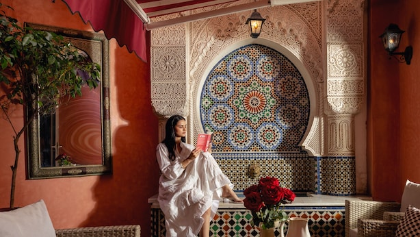 A woman reading in a room with ornate tile work, wicker furniture, and a decorative mirror.