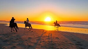 A beach scene with three people riding horses, silhouetted against a sunset.