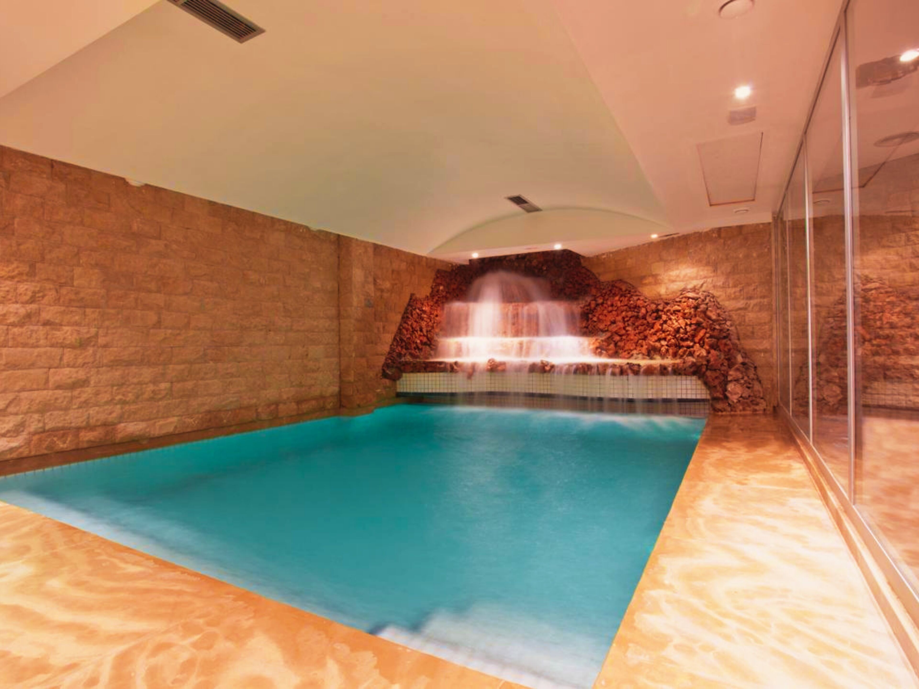 Una piscina interior con cascada, paredes de piedra y cerramiento de cristal.
