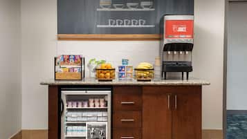 A hotel room with a kitchenette area featuring a refrigerator stocked with beverages, a coffee machine, and a fruit bowl.